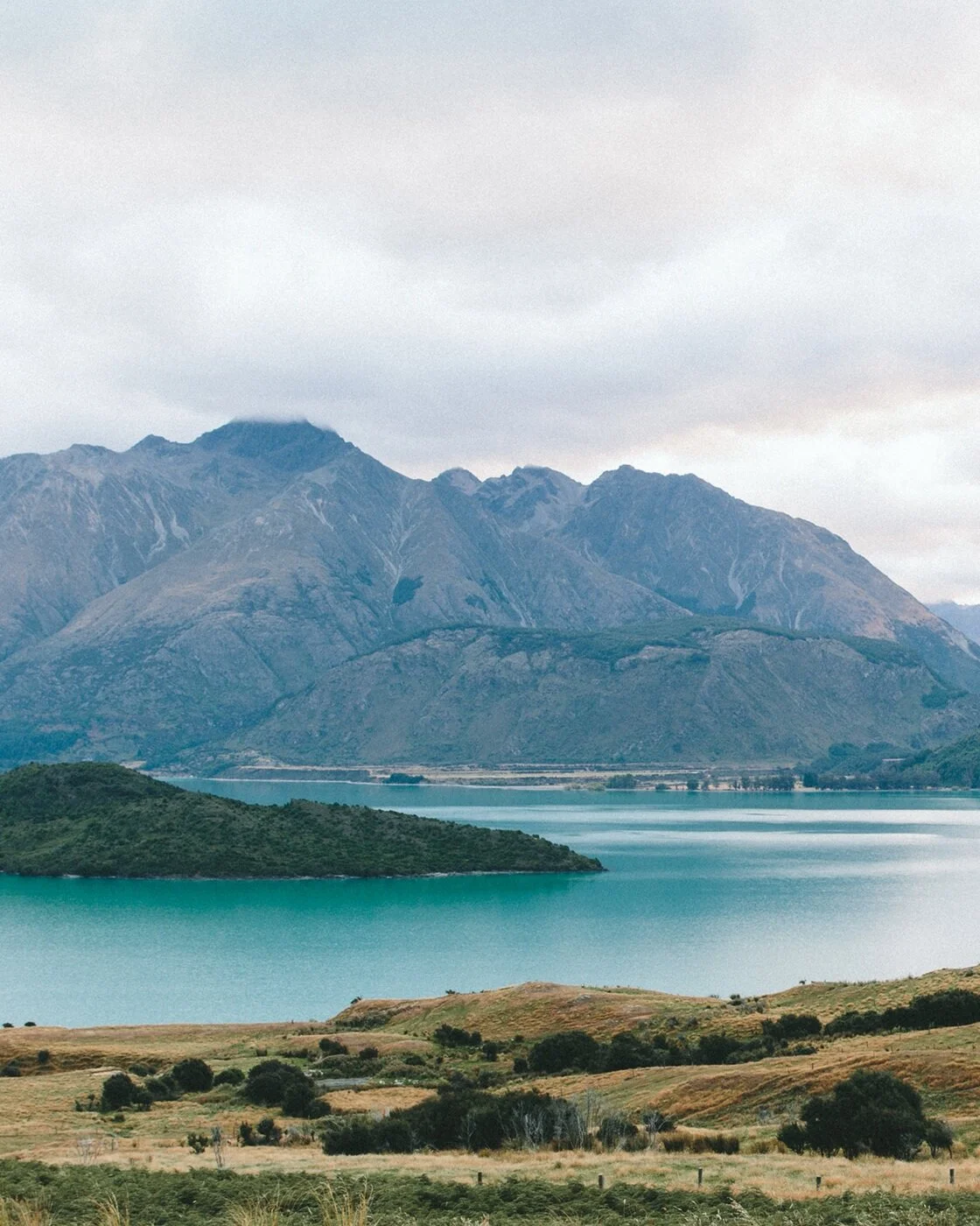 Scenic landscape of a large mountain range in the background, a lake in the middle with a small island, and rolling green hills in the foreground under a cloudy sky.
