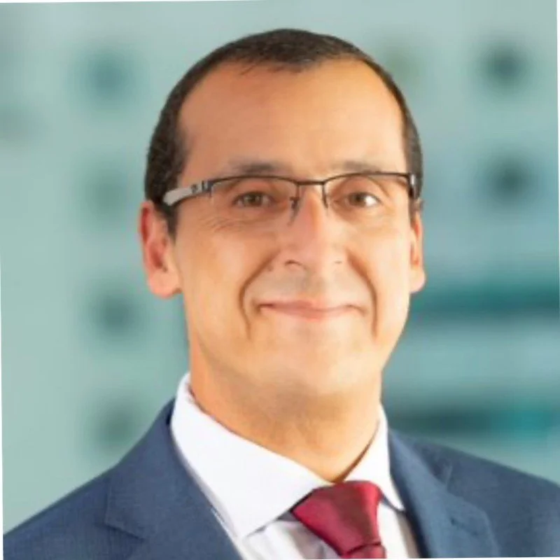Smiling person wearing glasses, a suit, and a red tie with a blurred background.