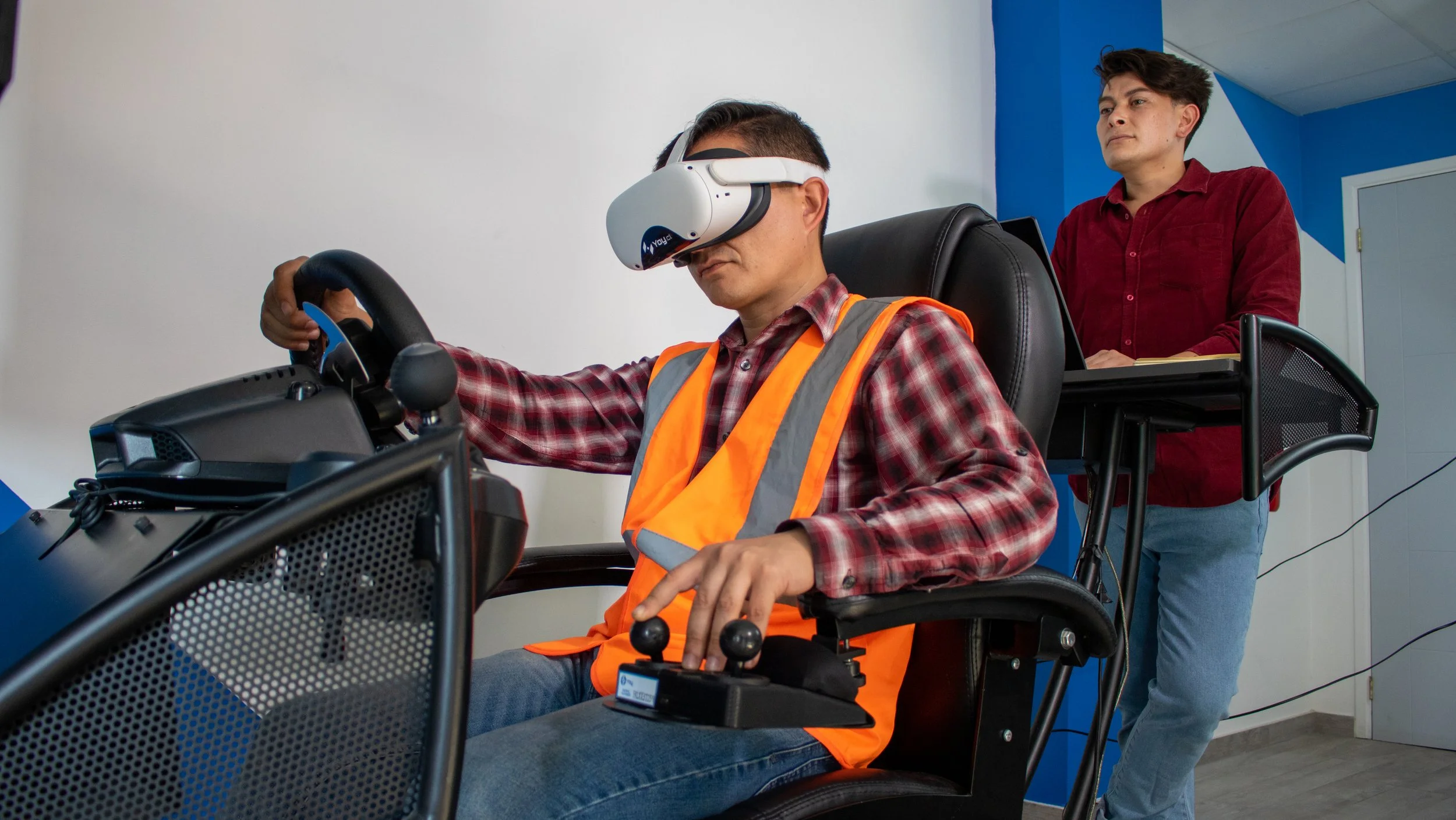 Person wearing a VR headset and orange safety vest using a simulator, while another individual observes.