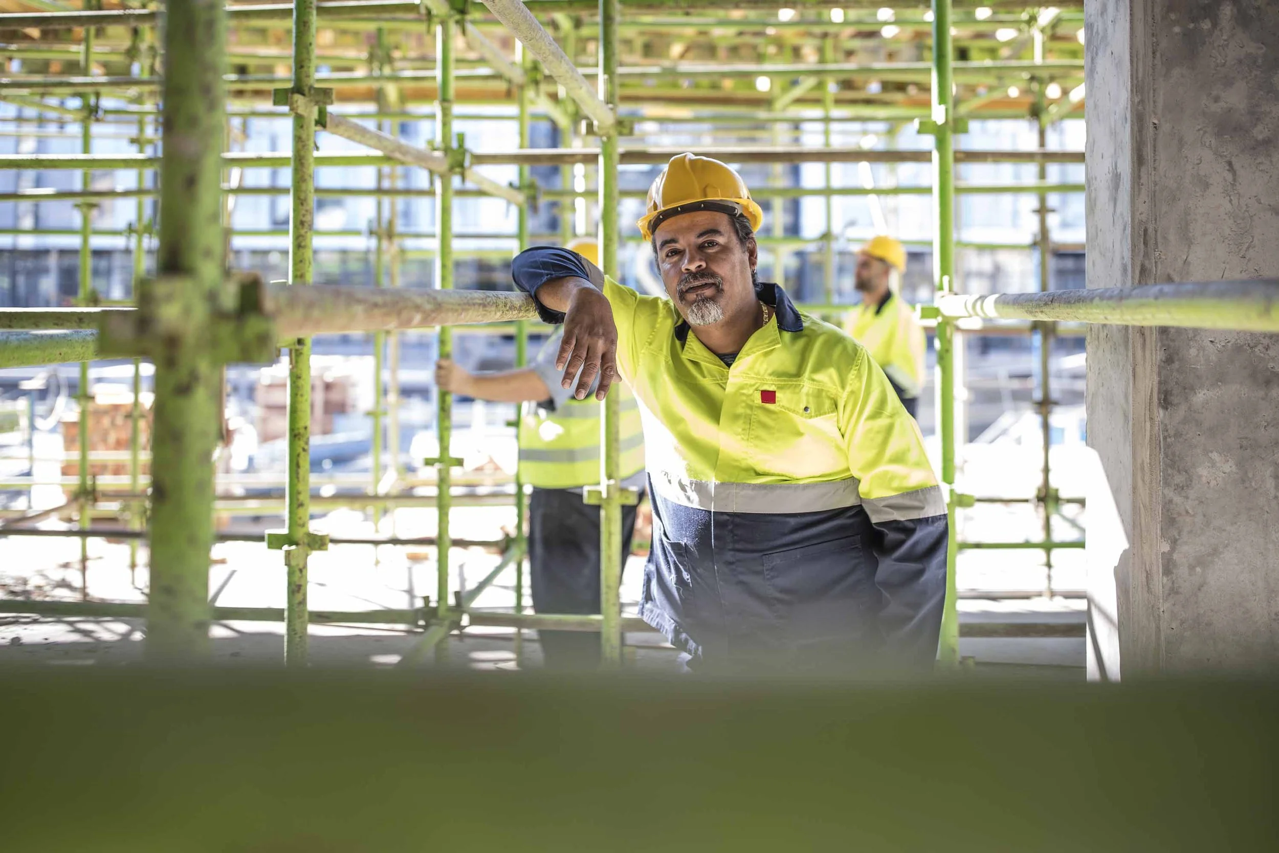 Construction worker in a yellow safety vest and helmet leaning on scaffolding at a building site, with other workers in the background.
