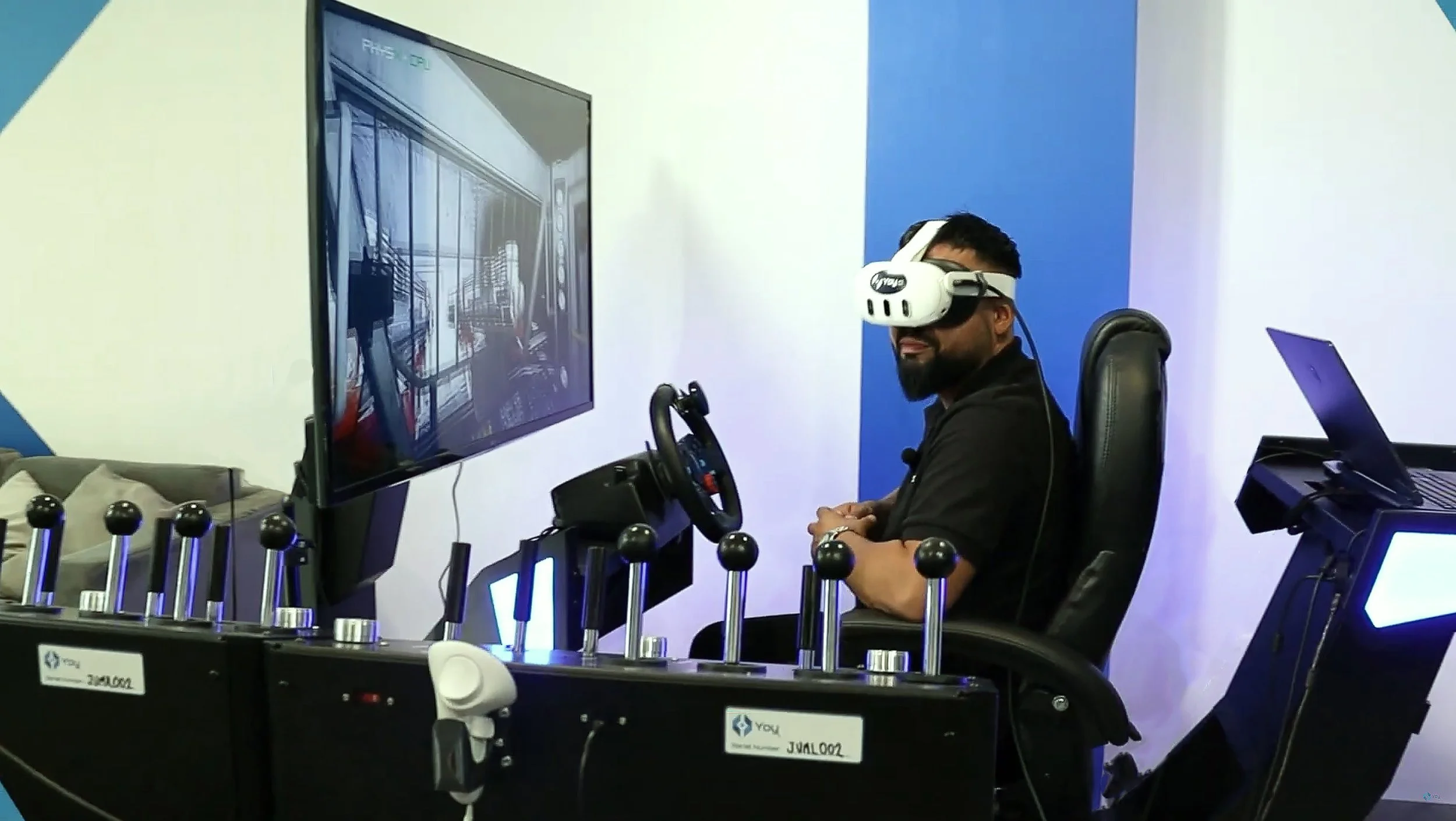 Hombre con visor de realidad virtual sentado en un simulador con una pantalla grande frente a él y un volante y pedales a su alrededor.