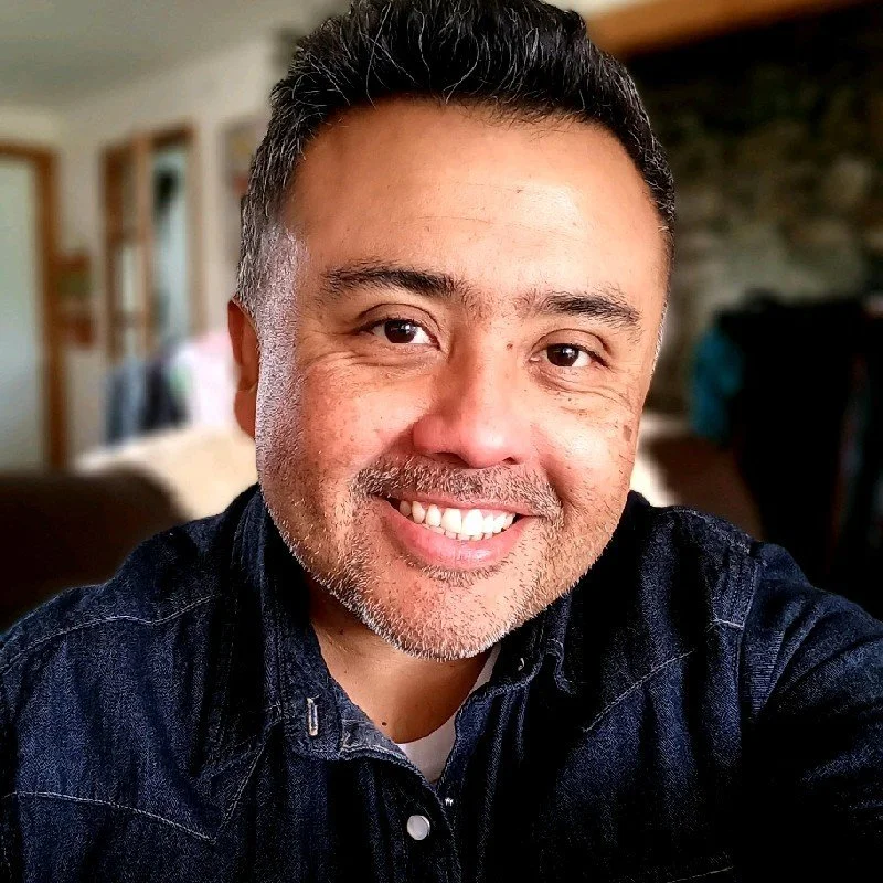 A person smiling at the camera indoors, wearing a denim shirt.