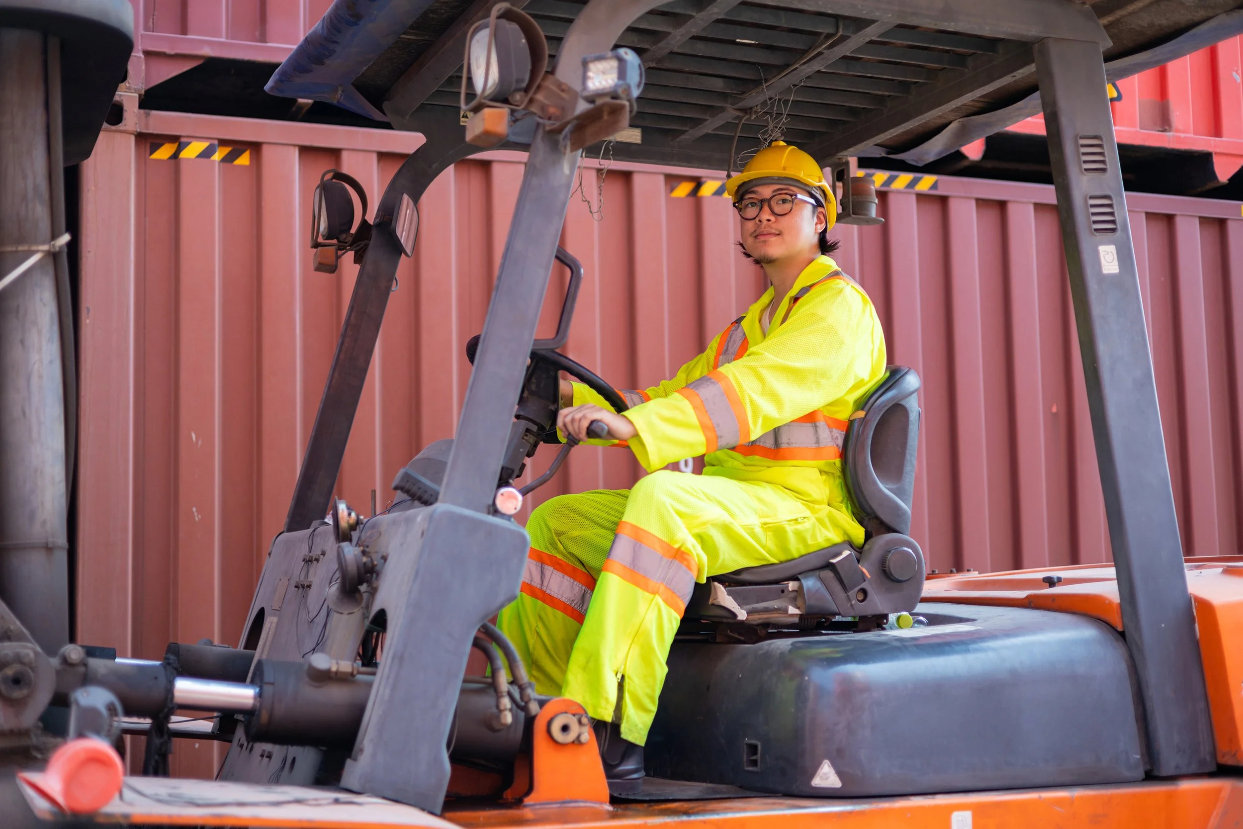A man wearing safety goggles, a yellow hard hat, and a neon yellow safety vest with orange reflective stripes is sitting and operating a forklift in front of a red shipping container.