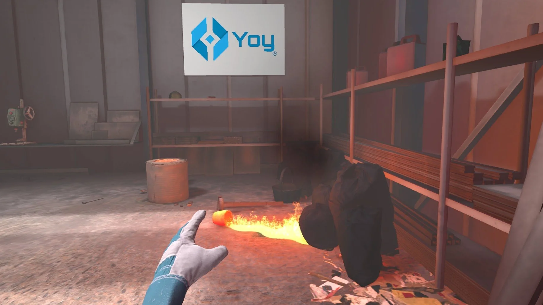 Escena de un juego de realidad virtual que muestra una mano con guante blanco señalando un balde derramado con llamas. El entorno parece ser una bodega industrial con estanterías con diversos artículos. En la pared hay un letrero con un logo azul y la palabra "yoy".