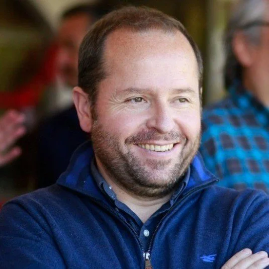 Smiling man in a blue sweater standing indoors with arms crossed.