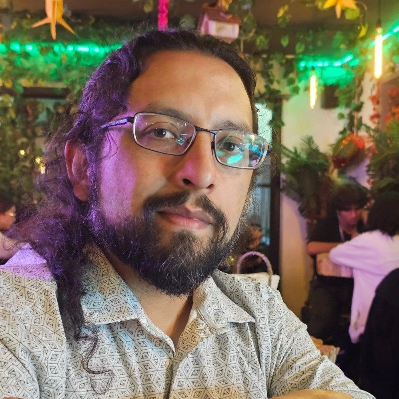 Victor Peñaloza, with glasses and a beard in a patterned shirt, sitting in a decorated indoor setting with plants and other people in the background.