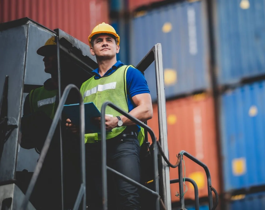 Trabajador de la construcción con casco y chaleco de seguridad cerca de contenedores de embarque, sosteniendo una carpeta.